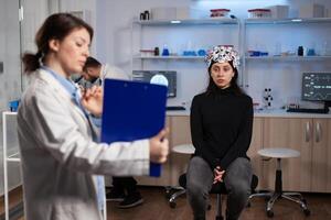 Patient woman wearing brain scanner standing on chair in neurology laboratory while neurologist doctor holding clipboard with tomography on it during neuroscience experiment. Nervous system diagnosis photo