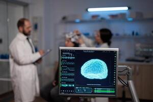Monitor with brain activity on screen in neurology medical laboratory. In background team of neurologist adjusting eeg headset of woman patient monitoring nervous system during neuroscience experiment photo