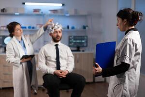 Researcher woman holding clipboard with neuroscience information on it while neurologist doctor adjusting eeg headset monitoring brain activity of man patient. Specialist analyzing nervous system photo