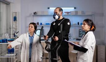 Medical team monitoring body endurance of sportman analyzing EKG data during medicine workout in modern laboratory. Athlete man with electrodes sensor on it running on fitness trainer photo