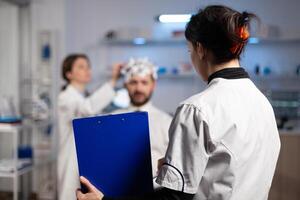 Doctor woman holding clipboard with neuroscience information on it while neurologist adjusting eeg headset monitoring nervous system of man patient. Physician engineer analyzing brain activity photo