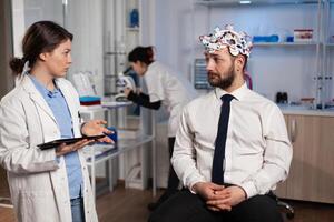 Neurologist woman doctor typing neurological disease symptoms on tablet computer discussing medical treatment with patient. Researcher engineer monitoring brain activity of man with eeg headset photo