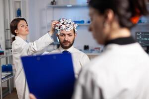 Specialist neurologist holding clipboard with explaining neurological treatment to man patient while researcher woman adjusting eeg headset. Physician doctor monitoring brain activity evolution photo