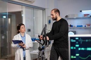 Runner man in sport equipment working at body endurance during medical examination in modern laboratory while woman researcher analyzing EKG data measuring heart rate pulse. Medicine service photo