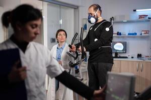 Specialist doctor looking at monitor analyzing heart rate pulse while athlete man running on gym bycle working at body endurance during medical workout in modern lab. Physician monitoring EKG data photo