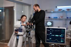 Specialist researcher doctor discussing with sportman in gym training equipment monitoring body endurance during medical workout in modern laboratory. Researcher doctor analyzing EKG data photo