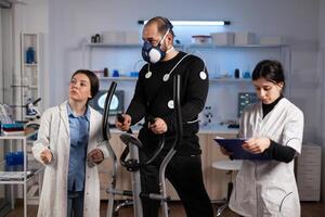 Specialist team analyzing body endurance of sportman during clinical workout in modern laboratory. Runner man with medical electrodes on it running on fitness trainer while doctor monitoring EKG data photo