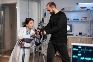 Specialist physician doctor talking with sportman in gym training equipment monitoring body endurance during medical workout in modern laboratory. Researcher doctor analyzing EKG data photo