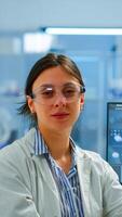 Chemist with lab coat sitting in laboratory looking at camera smiling, while colleague working on computer in background. Scientist examining virus evolution using high tech for scientific research photo