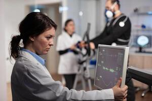 Scientist woman analyzing heart rate pulse on monitor while in background athlete man with electrodes working at body endurance running on fitness trainer in lab. Specialist doctor monitoring EGK data photo