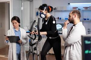 Athlete woman with mask running on gym bycle trainning body endurance while researcher doctor measuring heart rate monitoring EGK data in laboratory. Sportwoman with medical electrodes on it photo