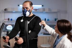 Specialist woman researcher putting electrodes sensor on sportman with mask monitoring EGK data in medical mordern laboratory. Athlete man running on fintess trainer working at body endurance photo