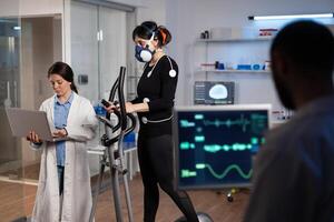 Athlete sportwoman with electrodes attached to her body running on cross trainer doing cardio exercise. Specialist researcher holding laptop analyzing endurance controling EGK data in modern lab photo