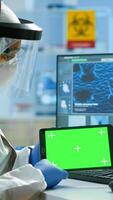 Doctor with coverall working at tablet with green screen in modern equipped lab. Team of microbiologists doing vaccine research writing on device with chroma key, isolated, mockup display. photo
