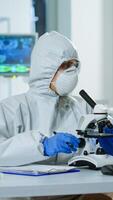 Medical research scientist conducting DNA experiments under microscope wearing protection suit and writing informations on clipboard. Chemist using high tech for vaccine development against covid19 photo