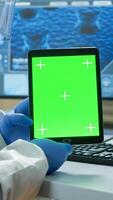 Chemist man wearing protection suit holding tablet with green mockup in modern equipped lab. Team of microbiologists doing vaccine research writing on device with chroma key, isolated display. photo