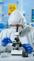 Scientist in ppe suit making adjustments and looking through laboratory microscope, typing on pc. Chemist in coverall working with various bacteria, tissue blood samples for antibiotics research photo