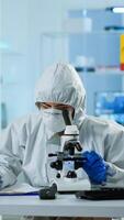 Biologist in ppe suit working on microscope and writing information on clipboard in equipped laboratory. Chemist examining vaccine evolution using high tech researching diagnosis against covid19 virus photo