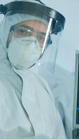 Microbiologist in ppe suit standing in laboratory looking at camera behind the glass wall in equipped lab. Doctor examining virus evolution using high tech and chemistry tools for vaccine development photo