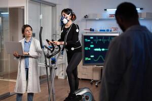 Specialist fitness researcher examining endurance controling EGK of athlete woman with mask running on bycle trainer. Physician man analyzing heart rate in medical modern laboratory. Medicine service photo
