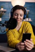 Portrait of social media influencer with headphones holding smartphone reading questions answering using production microphone sitting at desk in podcasting studio at home. On-air show photo