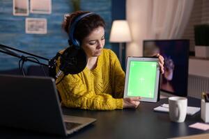 Social media creator with production headphones holding mock up green screen chroma key tablet with isolated display recording online podcast using livestream equipment. Vlogger streaming live content photo