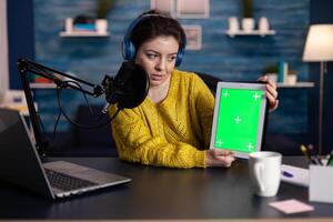 Social media influencer recording online for entertainment vlog holding mock up green screen chroma key tablet with isolated display in living room. Vlogger woman using podcasting equipment photo