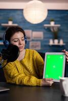 Vlogger woman recording livestream for entertainment vlog holding mock up green screen chroma key tablet with isolated display in living room. Social media influencer using podcasting equipment photo