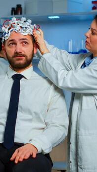 Woman researcher putting brainwave scanning headset to patient developing experiments using brain study lab monitors. Man siting on chair while scientist adjusting device examining nervous system photo