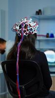 Back view of woman patient wearing performant eeg headset sitting on chair in neurological research laboratory while medical researcher adjusting it, examining nervous system typing on tablet. photo