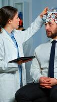 Man sitting on neurological chair with brainwave scanning headset, researcher doctor tracking evolution of patient's brain using tablet, adjusting eeg device. Scientist examining nervous system. photo