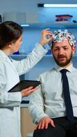Young man wearing brainwave scanning headset discussing with neurological researcher about symptoms of disease. Medical scientist writing informations of brain scan on tablet working in laboratory photo