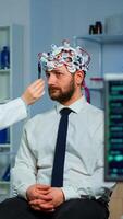 Man sitting on neurological chair with brainwave scanning headset while researcher examining health status writing on clipboard. Doctors working in brain study lab with monitors showing EEG reading. photo