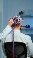 Back view of man patient wearing performant brainwave scanning headset sitting in neurological research laboratory while medical researcher adjusting it, examining nervous system typing on tablet. photo