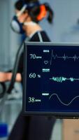 Medical researcher examining EKG image showing on monitor while patinet with mask running on cross trainer testing heart rate using electrodes.Doctor monitoring physical endurance in science sport lab photo