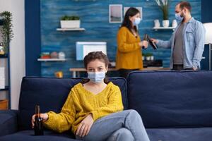 Lady looking at camera enoying spending time with friends in living room keeping social distancing wearing face mask to prevent coronavirus spreading. during global outbreak holding beer bottle. photo