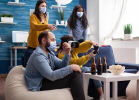 Young woman experiencing virtual reality playing games with vr headset and joystick in living room. friends drinking beer and laughing keeping social distancing because of global pandemic. photo