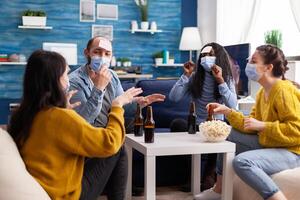 Mixed race of people with sticky notes on foreheads playing name game having fun with beer and popcorn keeping social distacing. with face mask to prevent spread of covid19 in home living room. photo