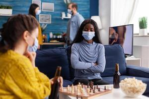 African woman wearing face mask playing chess drinking beer, keeping social distancing preventing spread of covid virus. Diverse people having fun in home living room playing board games photo