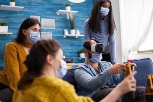 Group of multi ethnic friends having fun playing games using virtual reality headset and joystick wearing face mask keeping social distancing. Diverse people enjoying at new normal party. photo