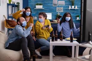 Group of multiethnic friends celebrating victory while playing games with joypad in living room keeping social distancing wearing face mask during global pandemic. People enjoying time together. photo