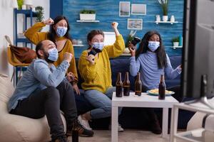 Excited multiracial gamer women sitting on couch playing games on console during new normal party wearing protective masks. Friends using wireless controllers keeping social distancing photo