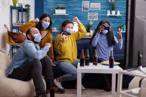 Multiethnic people celebrate game victory in home living room with joystick wearing face mask keeping social distancing in time of corona outbreak. Diverse friends enjoying beer and chips. photo