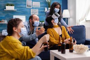 Man with face mask cheering up for friends playing games with vr headset in home living room keeping social distancing in time of global pandemic with covid. Multi ethnic people having fun. photo