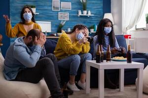 Disappointed diverse people loosing games at home respecting social distancing because of corona outbreak wearing face mask against spreading virus. New normal party social distance photo