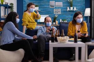 Diverse friends trying to win playing games using joystick havin fun wearing face mask to prevent spreading of coronavirus in time of global outbreak. Gaming competition, beer and popcorn. photo