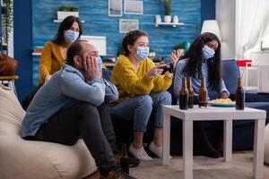 Sad multi ethnic group of friends after lose at gaming competiton with face covers with mask during covid outbreak keepin social distancing in home living room. Enjoying beer and chips. Conceptual image. photo