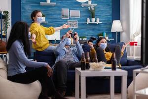 Multiethnic friends celebrating victory playing games in living room raising hands wearing face mask and keeping social distancing during global pandemic. People enjoying time together photo