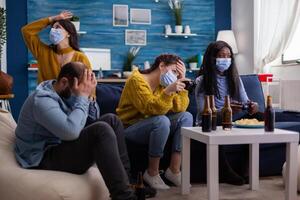 People loosing game playing at new normal party during social pandemic with face mask, keeping distancing against spreading covid 19 virus sitting on couch in living room photo