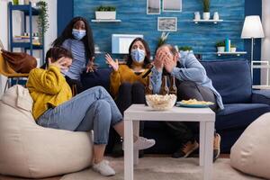 Group of multiethnic friends watching a horror movie on tv being afraid, spending weekend together wearing face mask to prevent infection with covid 19 during global pandemic enoying beer and chips photo